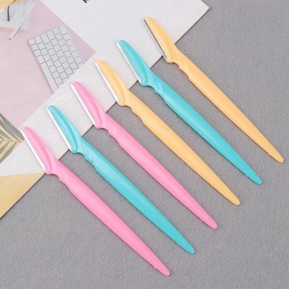 Zes gezichts-scheermesjes in pastelkleuren roze, turquoise en geel, op papier.